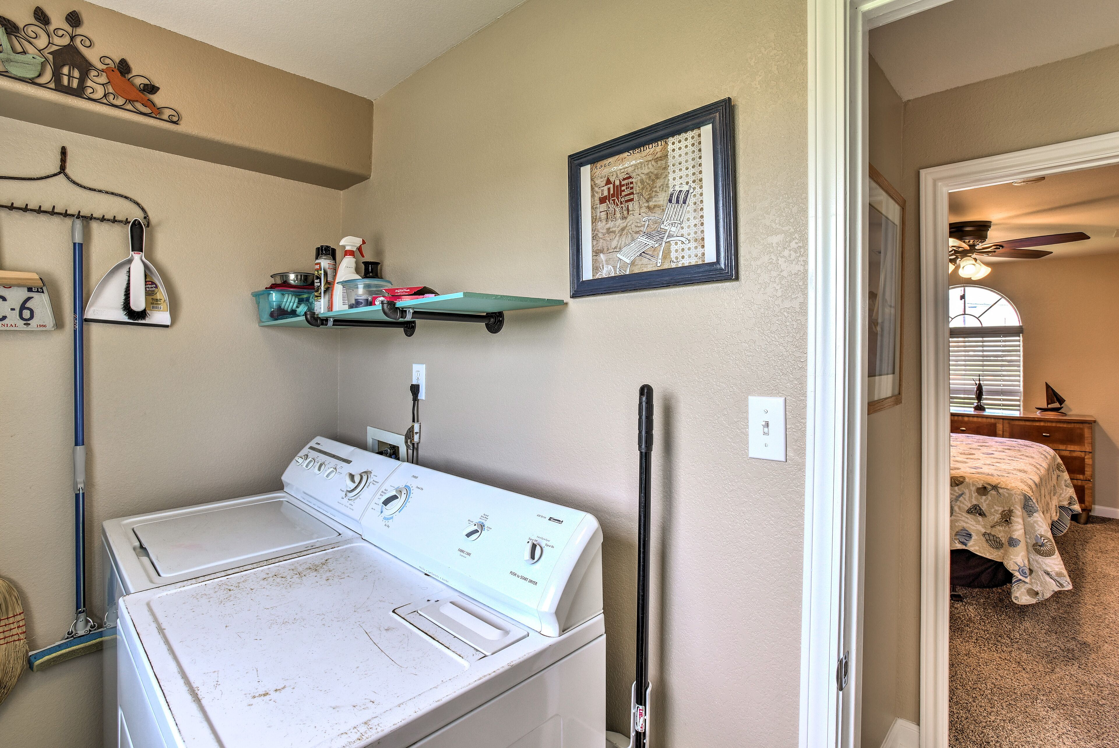 A laundry area with a front-loading washer and dryer, a broom, a shelf with cleaning supplies, and a framed picture on the wall.