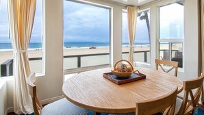 Une table ronde en bois, sur laquelle sont posés un panier tressé et un saladier, le tout mis en valeur par une vue sur la plage, visible à travers de grandes fenêtres.