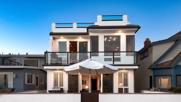Une maison moderne à deux étages, dotée d’un balcon, de grandes fenêtres et d’un espace extérieur aménagé et abrité.