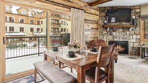 A wooden-floored dining area with a stone fireplace, a TV mounted above it, and a view of neighboring buildings through large windows. - Remodeled! Near Downtown Breckenridge! (Breckenridge)