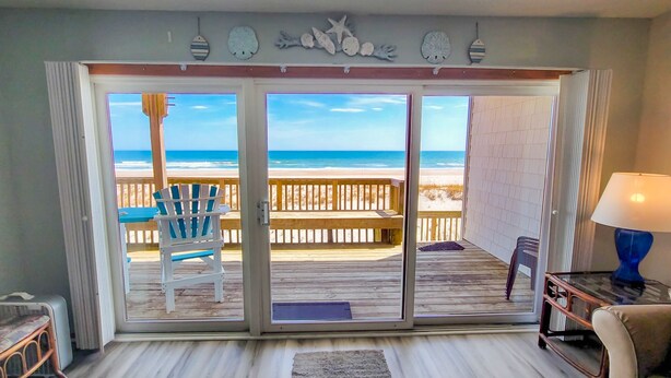 A room with a view of the beach, a wooden deck outside, and a sliding glass door. - Queen's Grant D-113 – Ocean Views & Beach Fun (Topsail Beach)