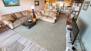 A living room with two beige sofas, a coffee table, a TV, and a dining area with a table and chairs. - Queen's Grant D-113 – Ocean Views & Beach Fun (Topsail Beach)