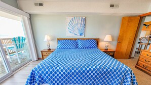 A bedroom with a large bed, two bedside tables with lamps, a wooden dresser, and a sliding glass door leading to an outdoor area with a chair. - Queen's Grant D-113 – Ocean Views & Beach Fun (Topsail Beach)