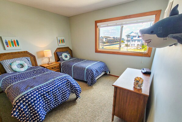 A hotel room with two beds, a wooden nightstand, a lamp, a window with a view of a house, and a shark decoration on the wall.