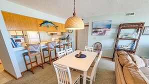 A dining area with a white table and chairs, a hanging fish decoration, a wooden ceiling, and a beach-themed wall art. - Queen's Grant D-113 – Ocean Views & Beach Fun (Topsail Beach)