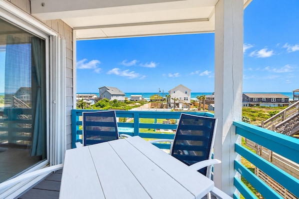 A balcony with a table and chairs, overlooking a beach with houses and a clear sky.