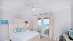 A bedroom with a bed, a ceiling fan, a TV, and a balcony with a view of the ocean.