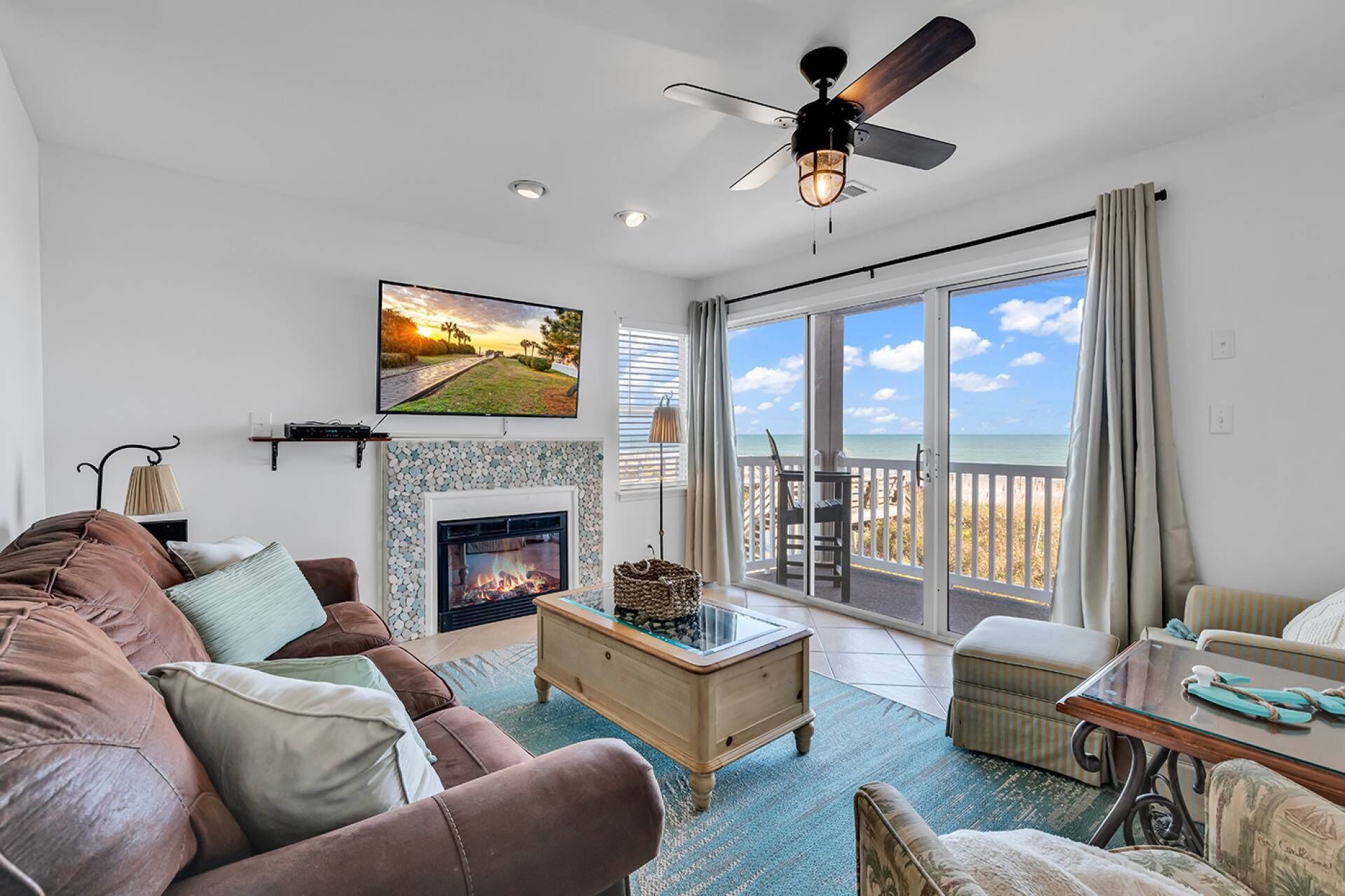 A living room with a fireplace, a sofa, a coffee table, and a view of the beach through large windows.