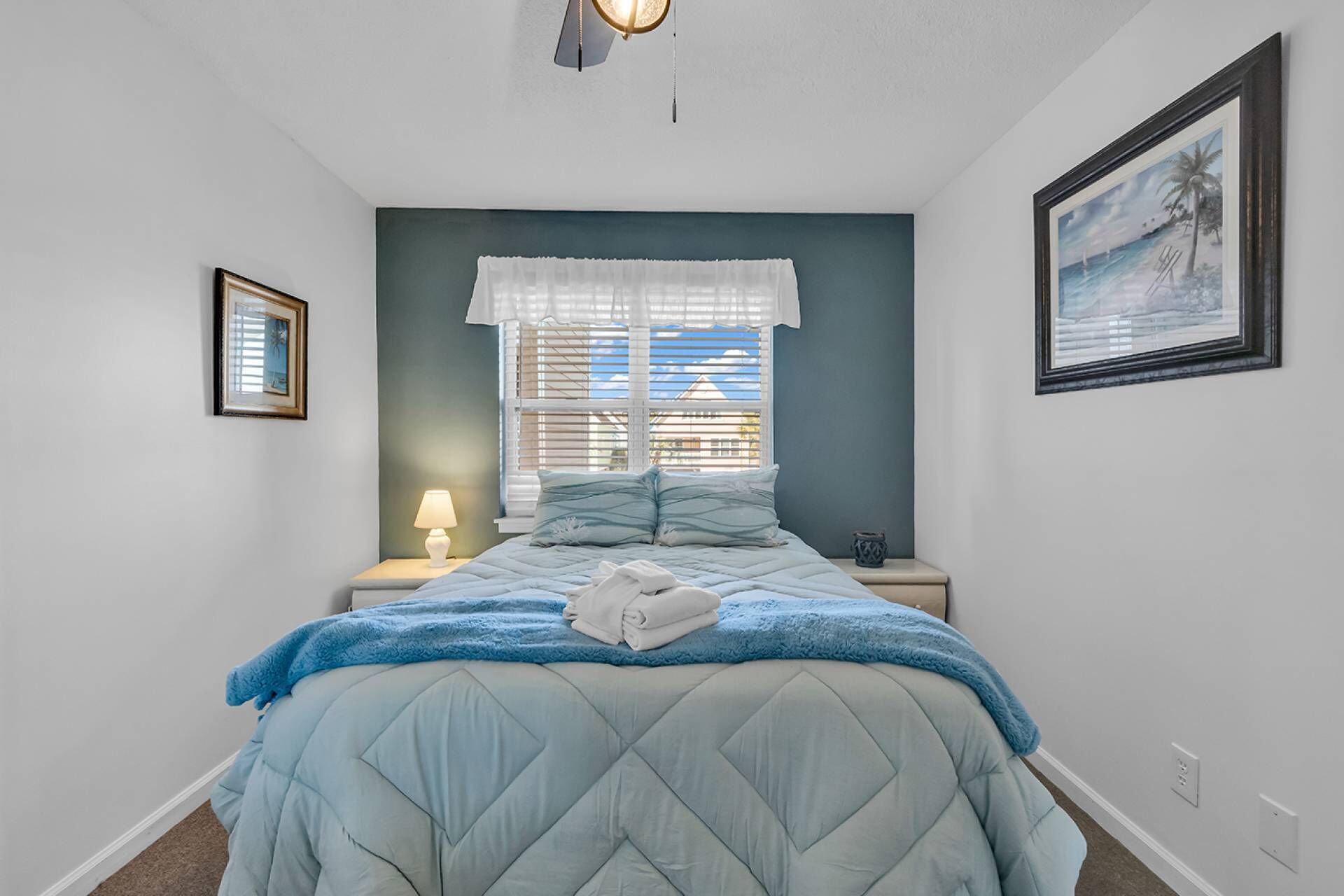 A bedroom with a bed, a nightstand, a lamp, a window with blinds, and a framed picture on the wall.