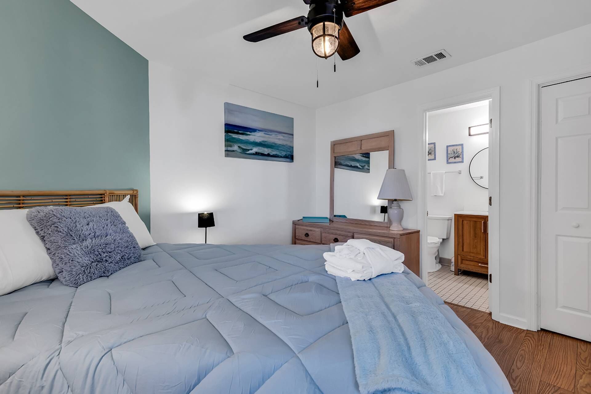 Une chambre à coucher comprenant un lit, un ventilateur de plafond, une table de chevet, un miroir et donnant sur la salle de bain.
