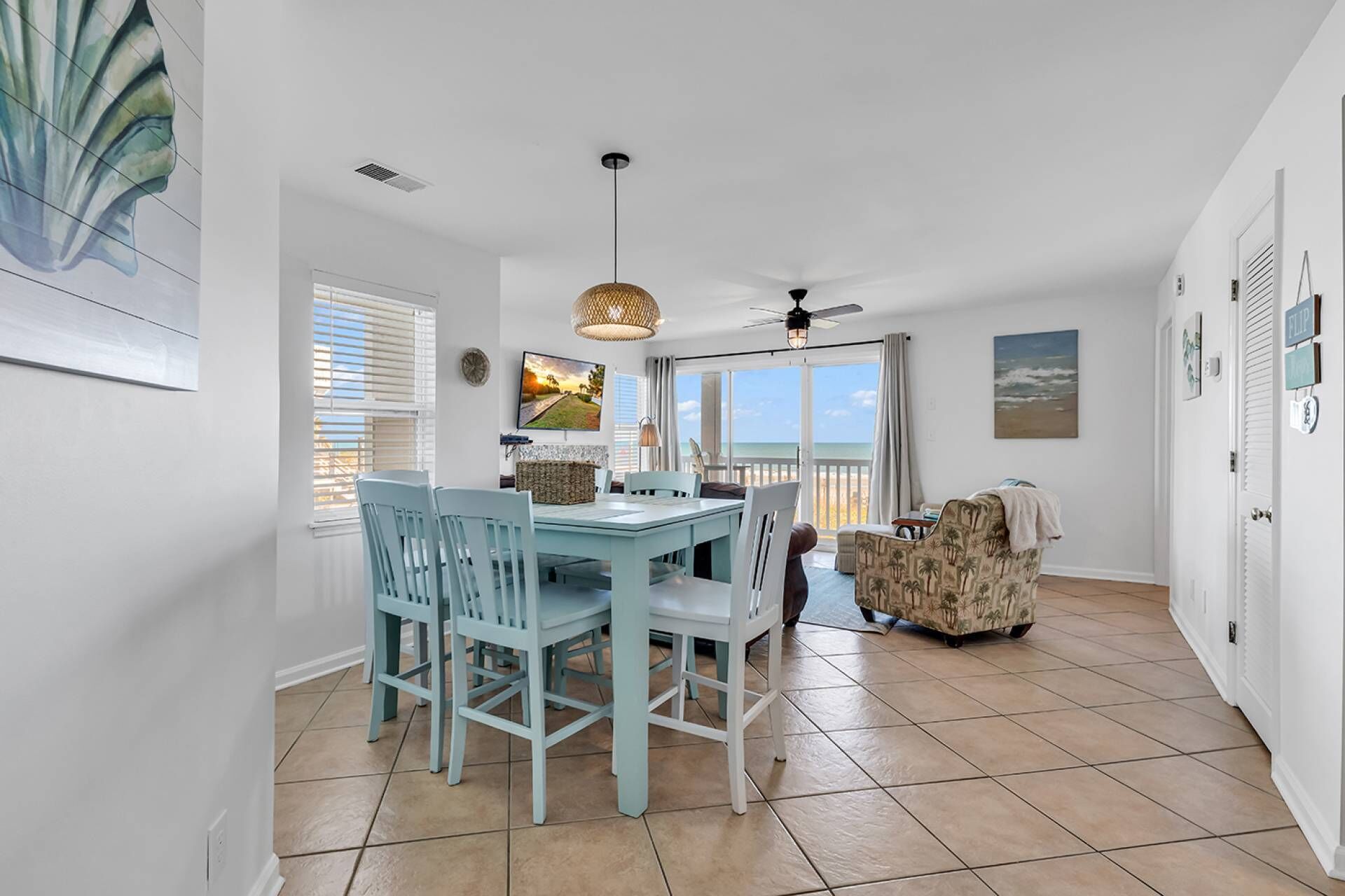A dining area with a table and chairs, a ceiling fan, and a view of the beach through large windows.