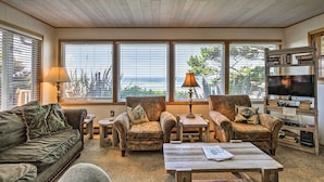 A cozy living room with a sofa, two armchairs, a coffee table, and a TV stand with books. Large windows offer a view of the outdoors. - Oceanfront Escape w/ Pacific Views: Surf & Explore (Arch Cape)