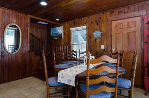 A wooden dining area with a table set for four, surrounded by wooden chairs with blue cushions. A staircase is visible in the background, leading to another level.