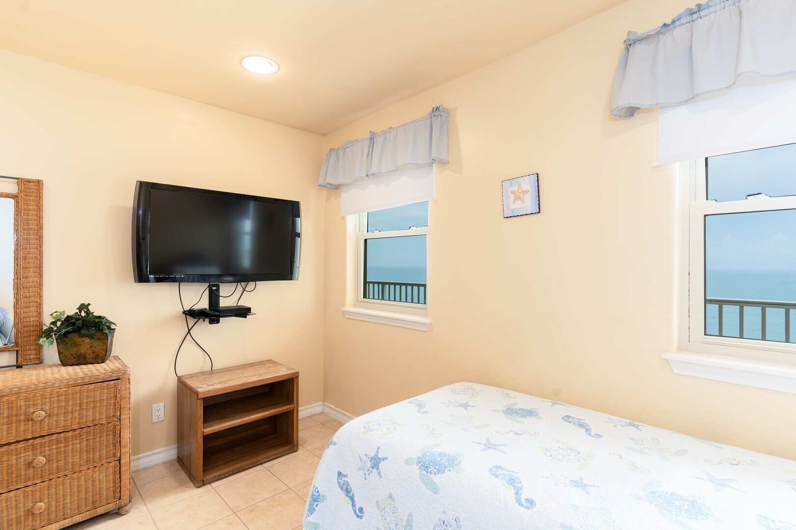 Un dormitorio con televisor montado en la pared, una pequeña estantería de madera, un aparador de mimbre, una cama con cubrecama estampado con estrellas y una ventana con vista al mar.