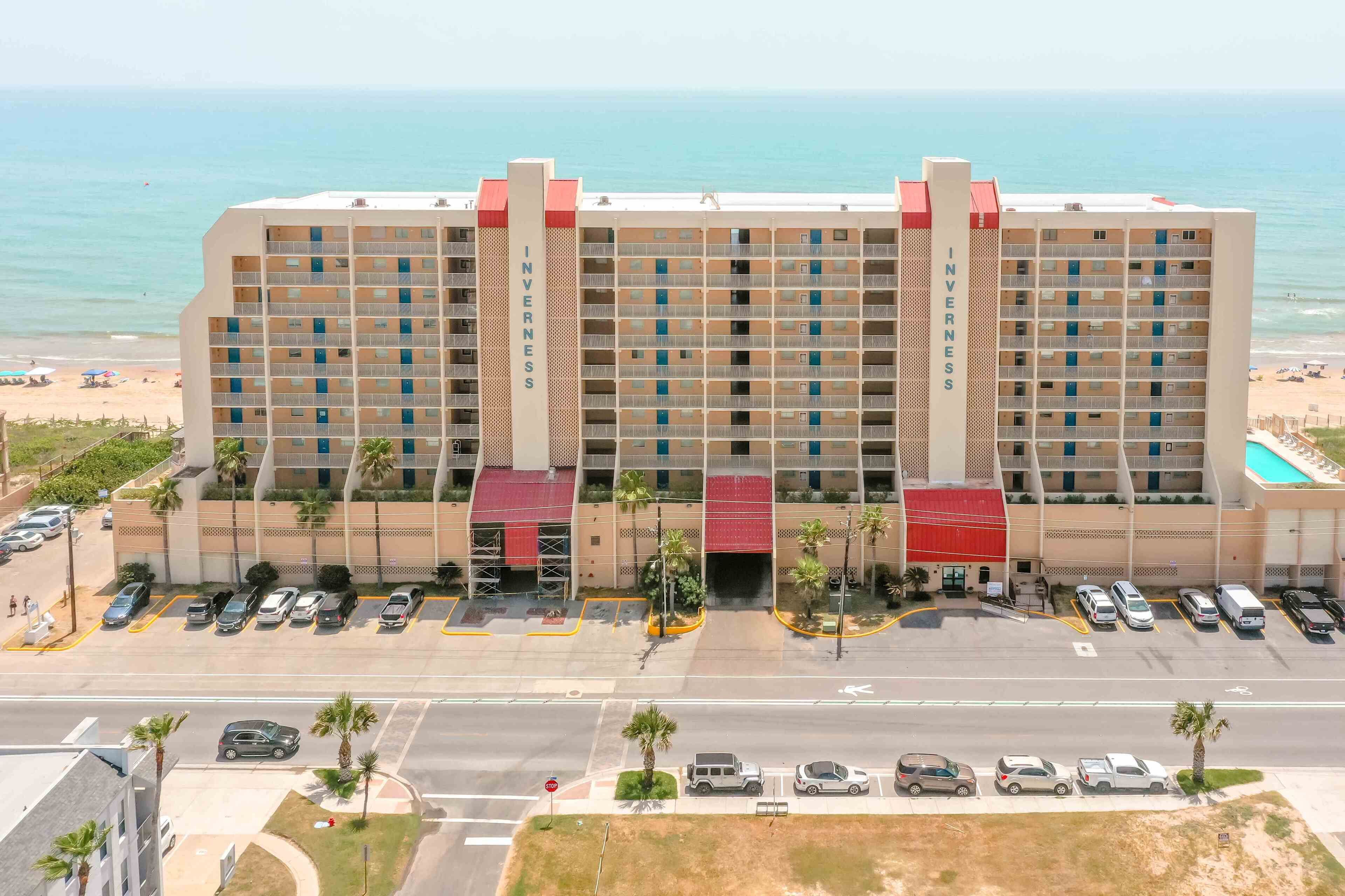 Un hotel de varios pisos con entrada central, estacionamiento y vista al mar.