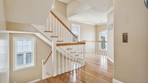 A spacious interior with a wooden staircase, white railings, and large windows allowing natural light. - Entire Home w/ pool, walk to beach (Virginia Beach)