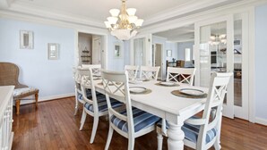A dining room with a white table set for six, surrounded by white chairs with blue cushions. A chandelier hangs from the ceiling, and the room has light blue walls and wooden flooring. - Entire Home w/ pool, walk to beach (Virginia Beach)