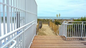 Una terraza de madera que conduce a una zona de playa arenosa, con una valla blanca y vistas al océano.