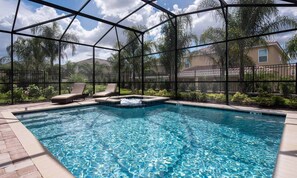 Una piscina con un jacuzzi, rodeada de sillas de descanso y una valla metálica, con casas y árboles al fondo.
