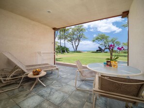 A patio area with two lounge chairs, a small table with a bowl of fruit, and a glass table with a vase of flowers. - Aloha Aku Hula Suite – Find Peace by the Shore (Kihei)