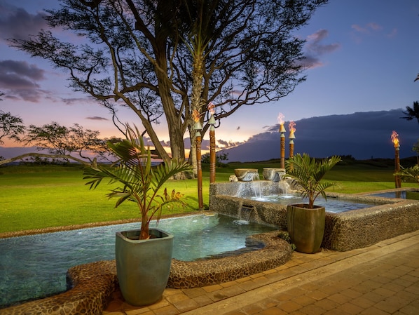 A serene outdoor pool area with a waterfall, surrounded by tropical plants and a large tree.