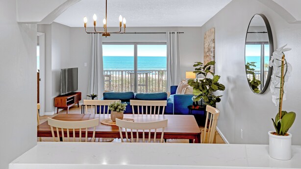 Een moderne woonkamer met een eetgedeelte, grote ramen met uitzicht op zee, een kroonluchter en een potplant.