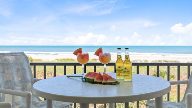 A round table with two glasses of a pink drink, a watermelon slice, and two bottles of a yellow beverage, set against a beach view. - Seaside Luxury Private Balcony, Breathtaking Views & Steps to the Sand (Cape Canaveral)