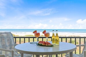 Een ronde tafel met twee glazen met een roze drankje, een plak watermeloen en twee flessen met een geel drankje, met uitzicht op het strand.