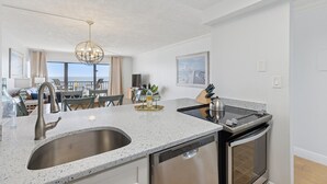 Een moderne keuken met een roestvrijstalen spoelbak, een vaatwasser en een oven. Achtergrond toont een eethoek met een tafel en stoelen.