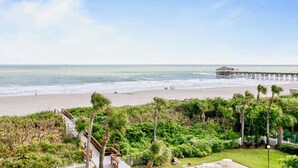 A beach view with a wooden pier extending into the ocean, a grassy area with palm trees, and a white fence. - Penthouse Paradise Steps to Pier with Ocean Views & Private Beach Access (Cocoa Beach)