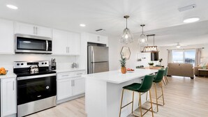A modern kitchen with white cabinetry, stainless steel appliances, a central island with bar stools, and pendant lighting. - Penthouse Paradise Steps to Pier with Ocean Views & Private Beach Access (Cocoa Beach)