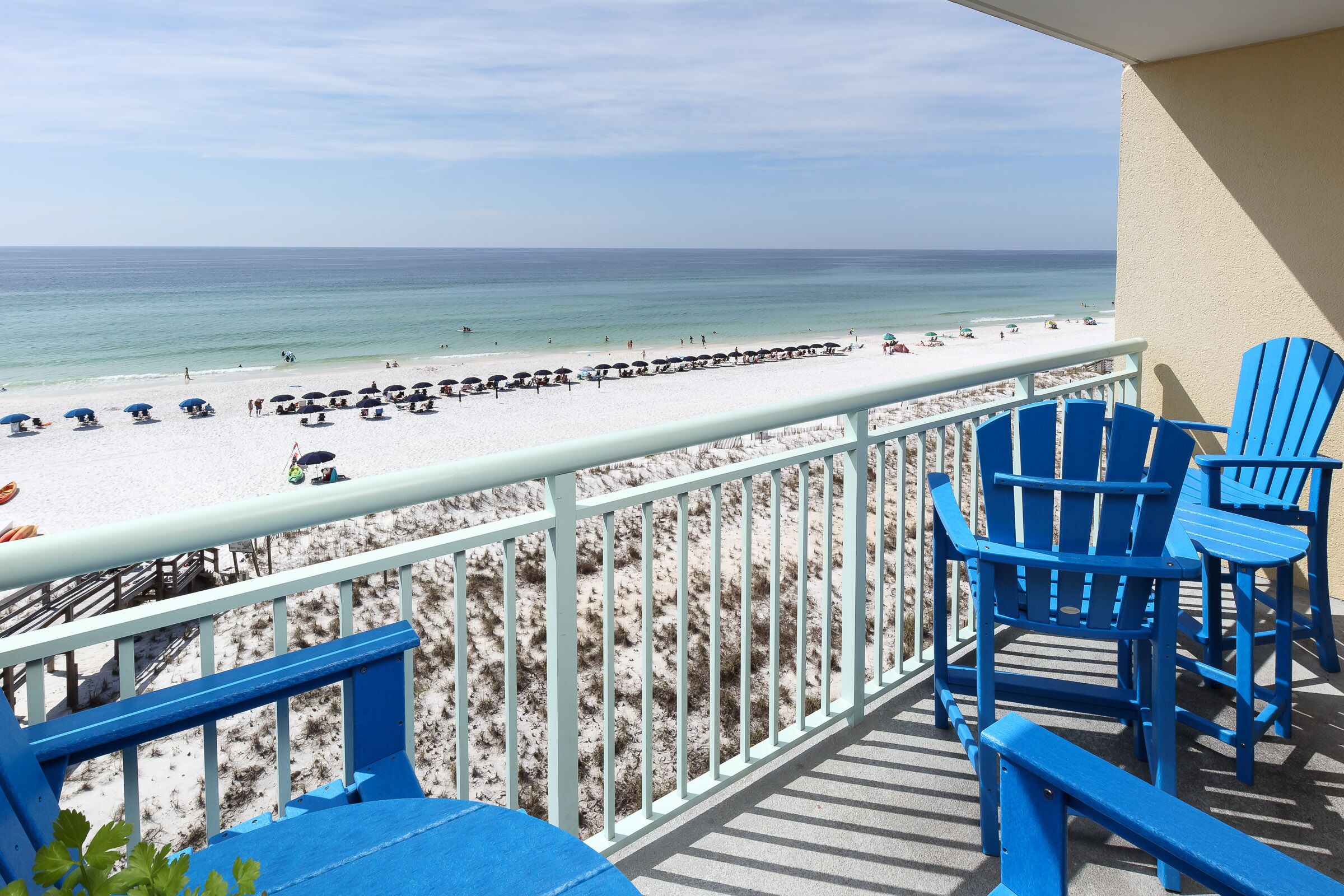 A balcony with blue Adirondack chairs overlooking a beach with umbrellas and people.
