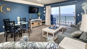 Una sala de estar con comedor, un ventilador de techo, un televisor montado en la pared y un amplio ventanal con vista a la playa.