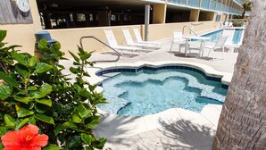 Un área de piscina con sillones de descanso y un jacuzzi, rodeado de follaje y un edificio con balcones.