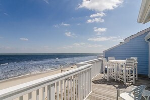 Une terrasse en bois avec des chaises blanches et une table, donnant sur une plage où l’on peut voir les vagues.