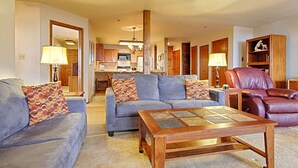 A living room with a sofa, a coffee table, and a leather armchair. There is a kitchen in the background.