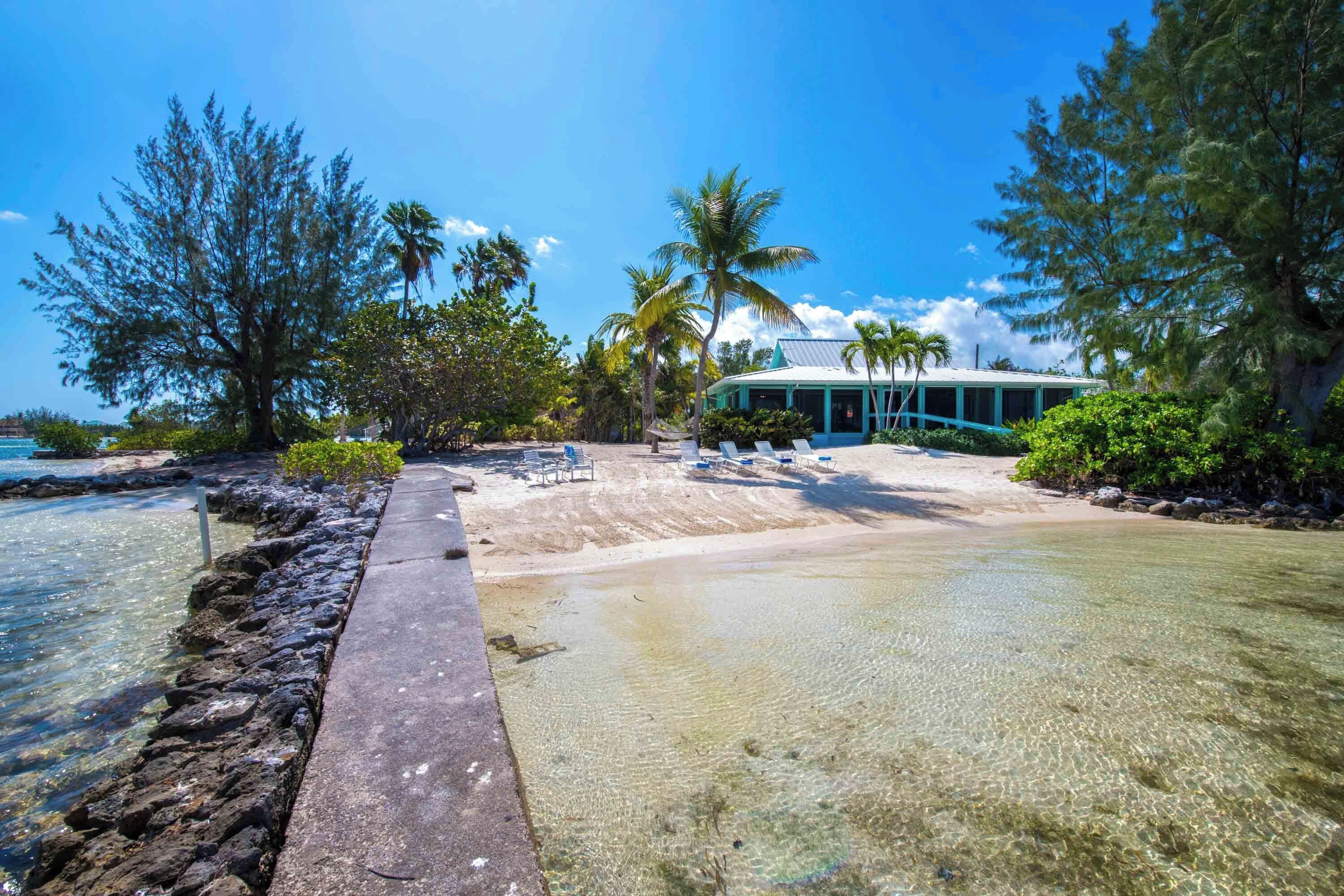 A beachfront property with clear water, a concrete walkway, lounge chairs, and a building with a green roof.