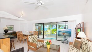 A spacious living area with a dining set, a sofa, a coffee table with flowers, a TV, and a view of the ocean through large glass doors. - Fingertip: Gorgeous Waterfront Villa in Cayman Kai w/Private Beachfront! (Grand Cayman)