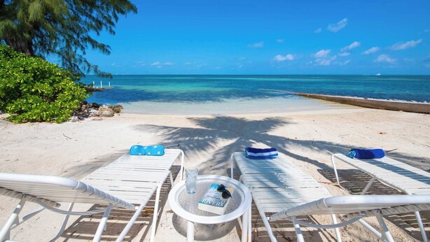 A beach view with lounge chairs, a table with a book and sunglasses, and a clear blue sky. - Fingertip: Gorgeous Waterfront Villa in Cayman Kai w/Private Beachfront! (Grand Cayman)