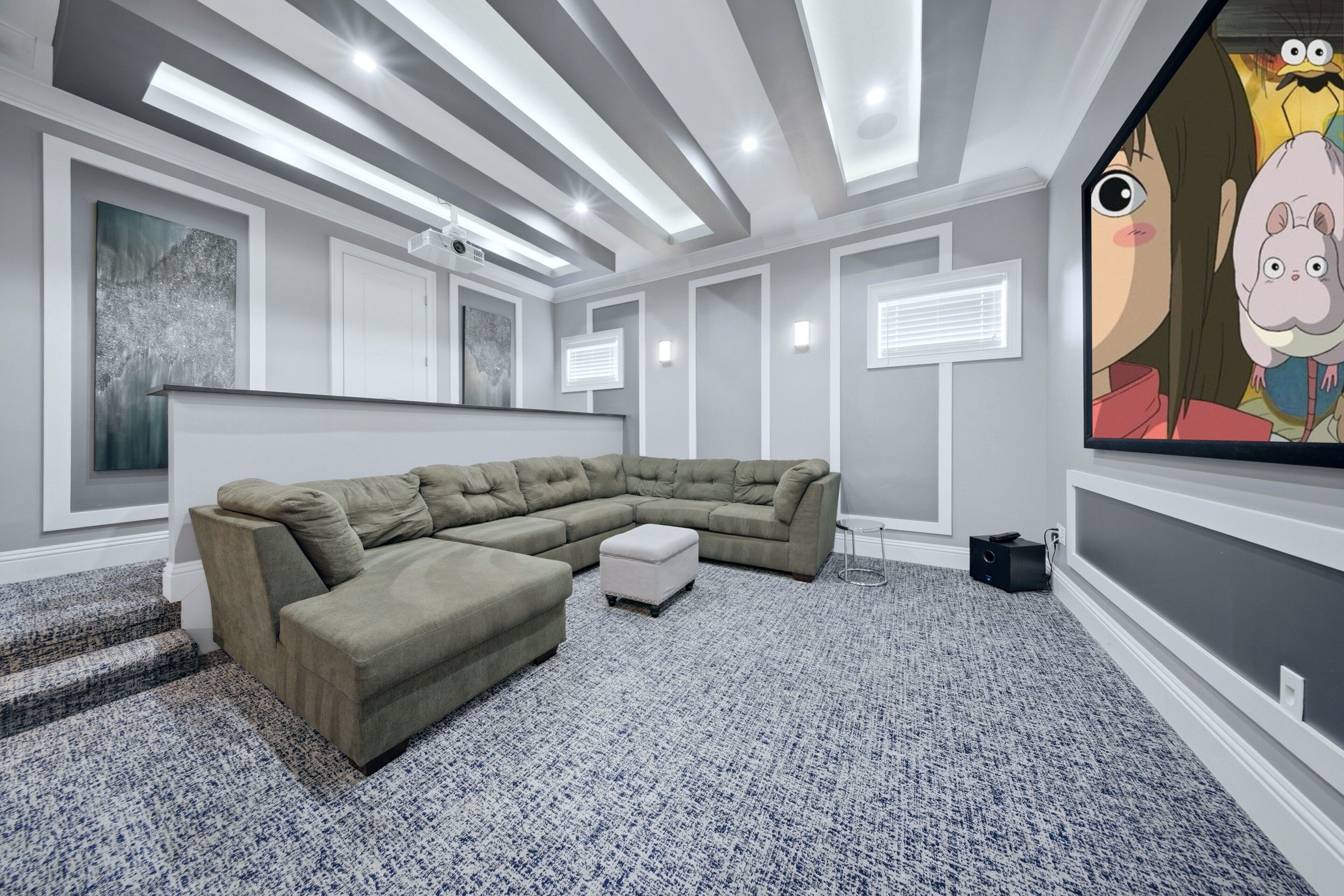 Un salon moderne avec un canapé d’angle, un petit pouf, un téléviseur à écran plat et un projecteur.