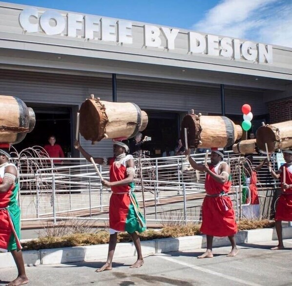 Un groupe de personnes vêtues de rouge et de vert se produit avec de gros troncs d’arbres devant un bâtiment sur lequel est affiché un panneau indiquant « CAFÉ – CRÉATIONS ORIGINALES ».