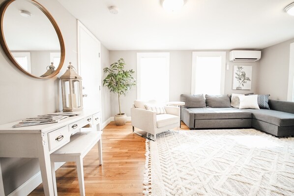 A modern living room with a white desk, a grey sofa, a white armchair, a large mirror, and a potted plant.