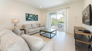 Ein modernes Wohnzimmer mit einer Couch, einem Couchtisch und einem Fernseher. Ein Fenster mit Vorhängen bietet einen Blick ins Freie.