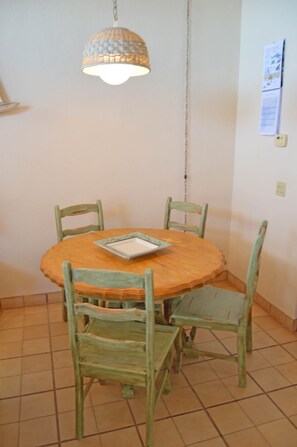 A round wooden dining table with a square placemat, surrounded by four matching chairs. A hanging lamp with a woven shade is above the table.