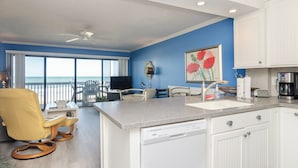 A modern kitchen with white cabinets, a granite countertop, and a dishwasher. There is a dining area with a yellow chair and a table, and a large window offering a view of the ocean.