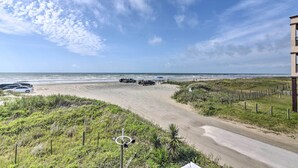 A beach view with a road curving through a grassy area, leading to the beach. - Furnished Deck, Beachfront Fun! Galveston Getaway (Galveston)