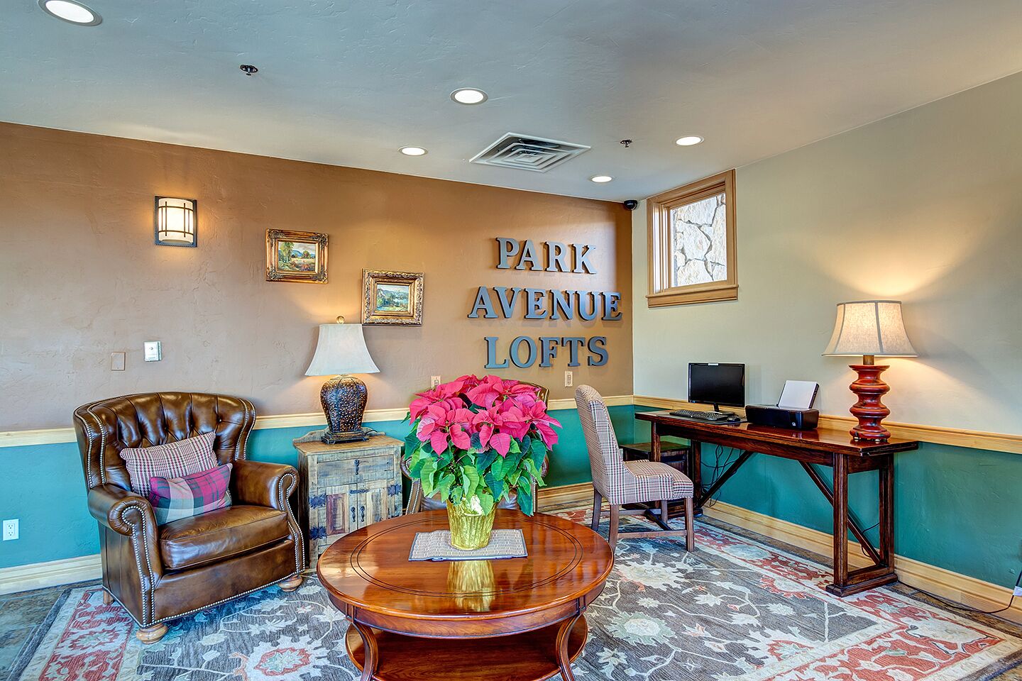 En lobby med en læderlænestol i brunt, et træbord med en vase blomster, et skrivebord med en computer, og et skilt på væggen der siger 'PARK AVENUE LOFTS'.