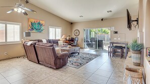 A spacious living room with a brown leather sofa, a coffee table, and a ceiling fan. There is a sliding glass door leading to an outdoor patio area with a table and chairs. - Heated Pool at 3 BR Home: 5 Mi to Coachella! (La Quinta)