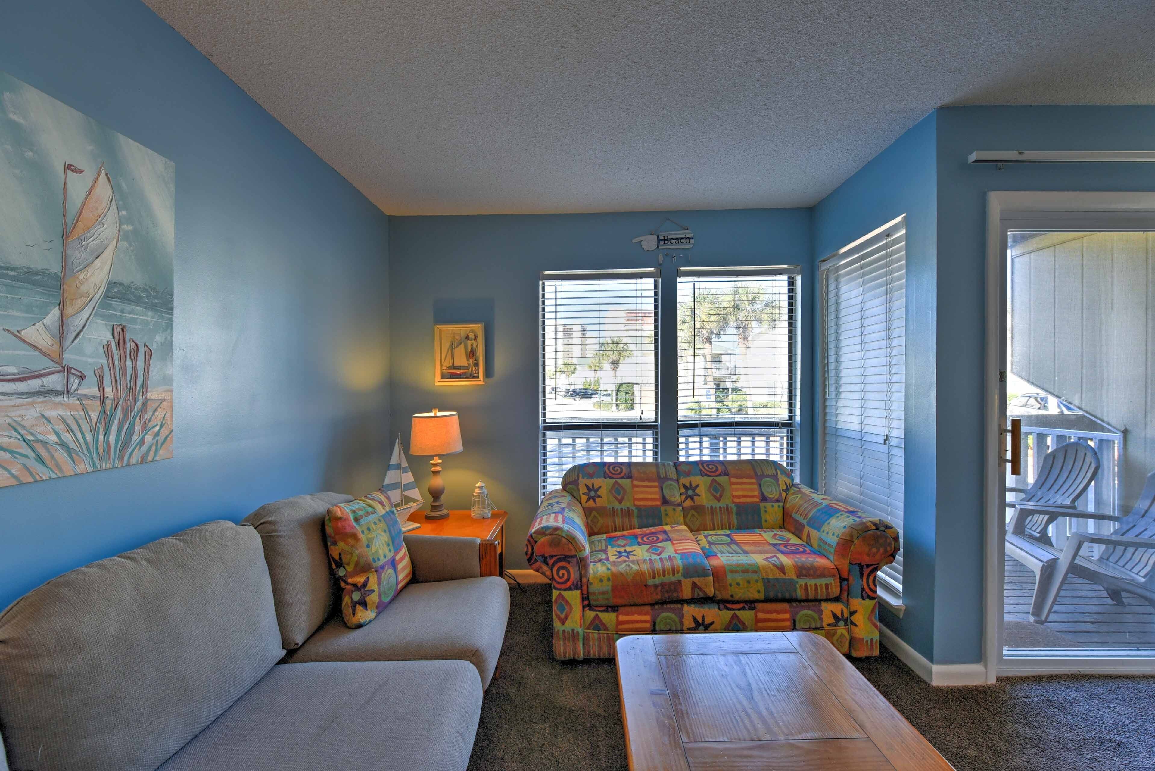 Ein Wohnzimmer mit blauer Wand, einer Couch, einem farbenfrohen Sessel, einem Holztisch und einem Fenster mit Jalousien.