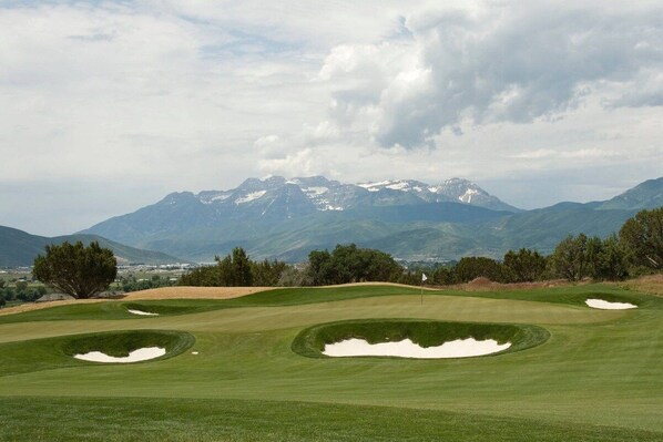 A golf course with bunkers and a flagstick, surrounded by mountains and trees.
